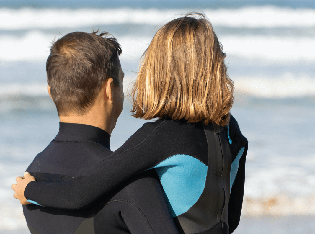 Father and daughter wear wet suits and stare at the ocean.