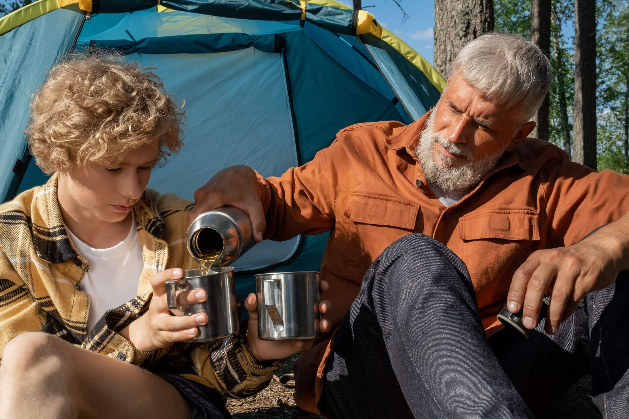 Grandfather pours grandson a cup of tea outside camping tent.