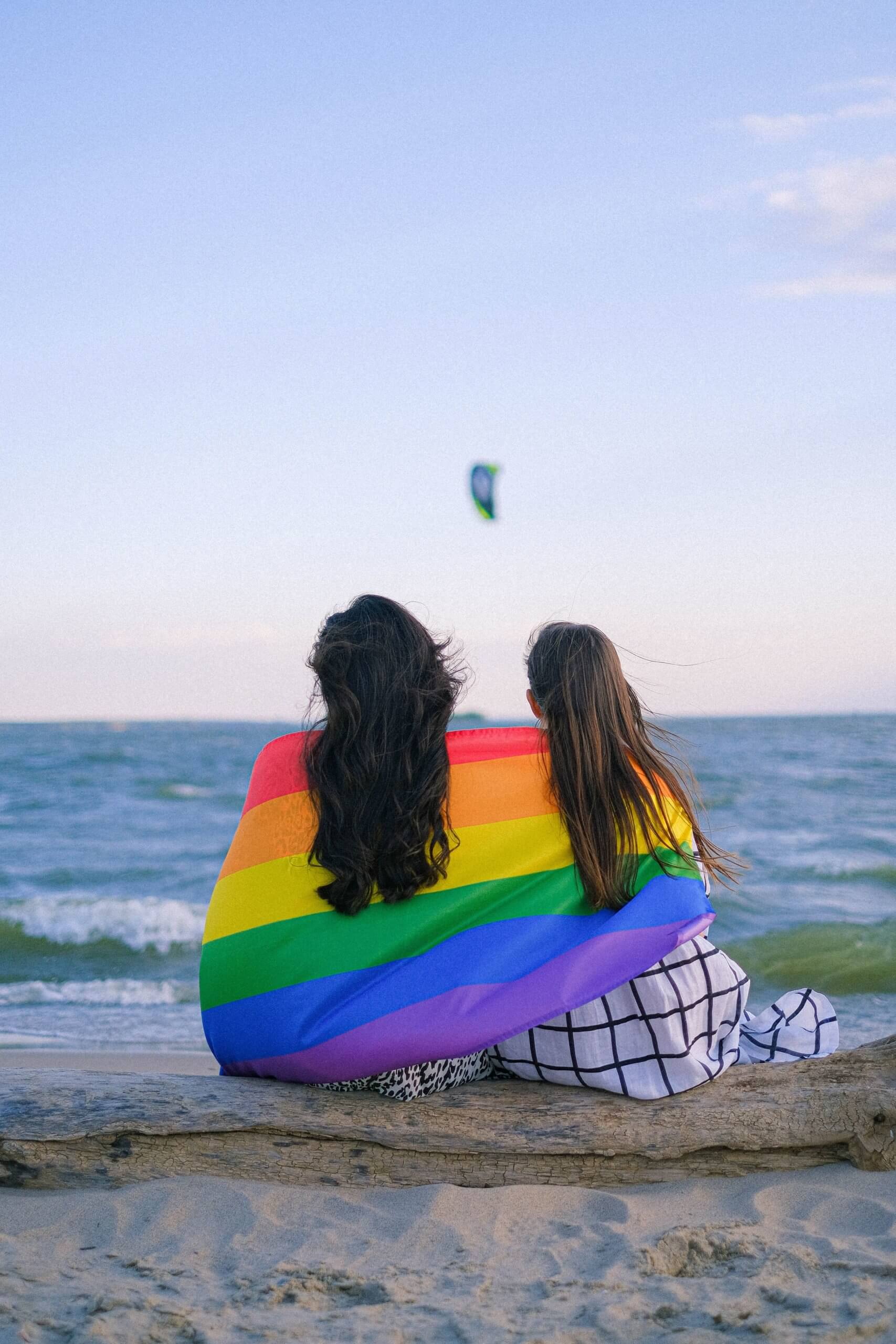 Two people sit on the beach wrapped in a Pride flag.