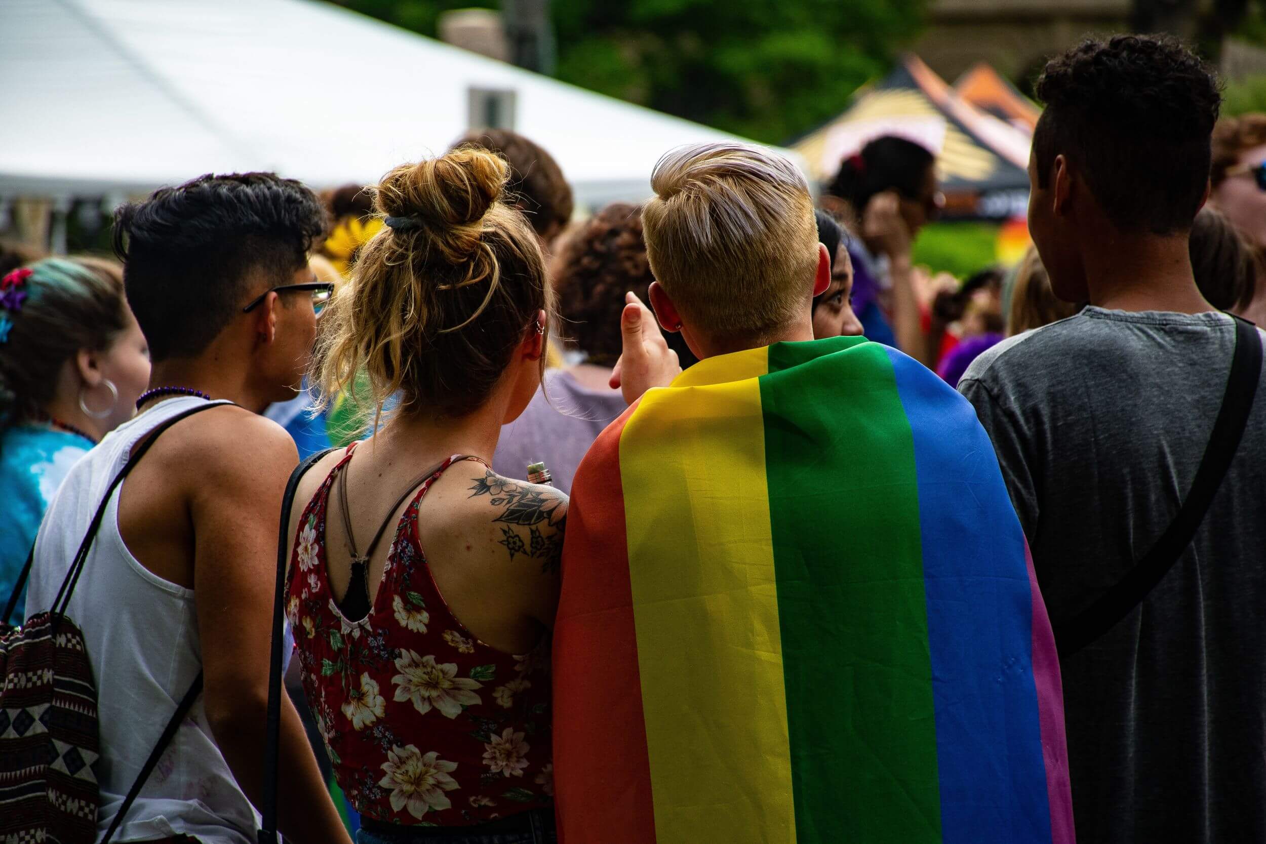 Four people stand at an outdoor event with their backs tuned. One of them has a pride flag draped around their shoulders.